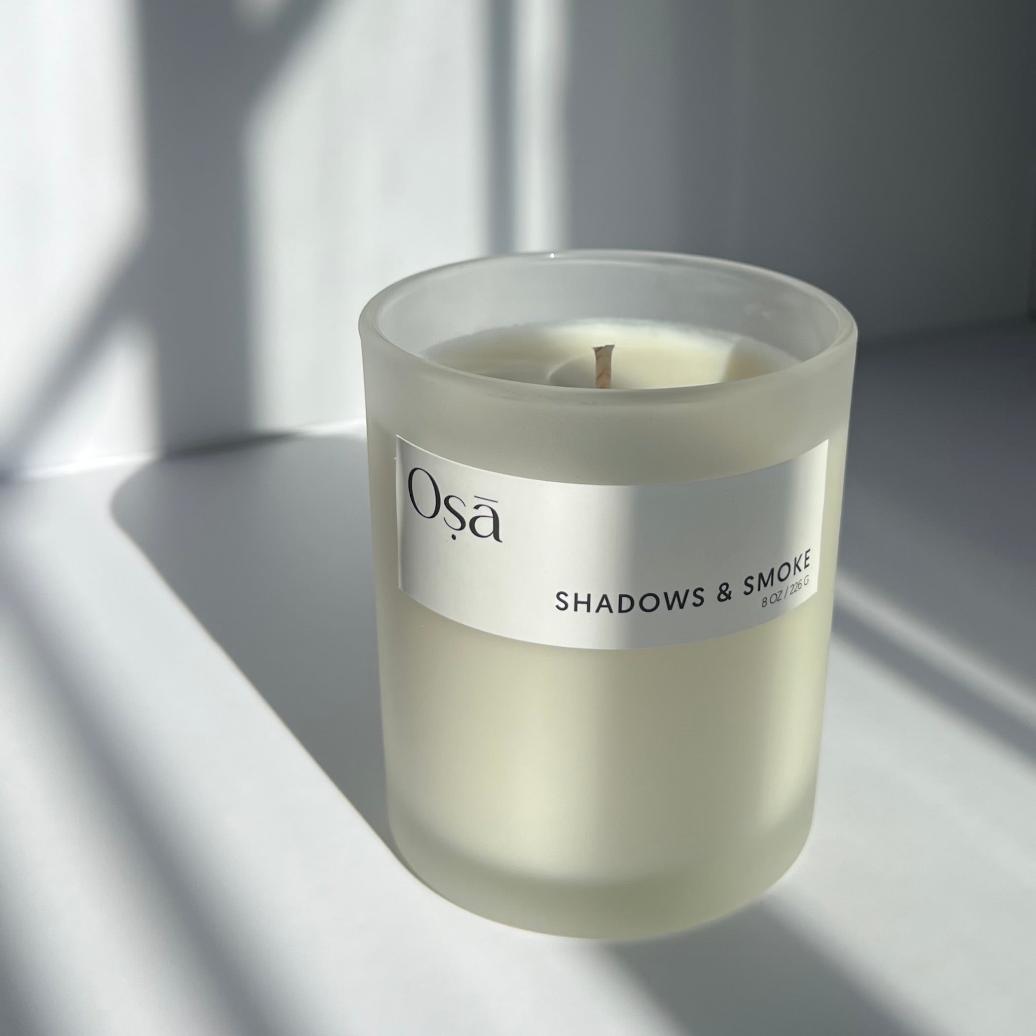 A candle in the sunlight with a label that displays Osa Shadows & Smoke.